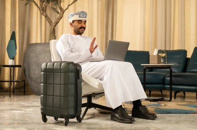 اجراء مكالمة فيديو ، رفع كف اليد بتعابير التحية و السلام، الجلوس في إحدى الفنادق الراقية، العمل عبر الحاسب المحمول، استخدام التقنيات الحديثة والمتطورة، رجل عربي خليجي عماني يرتدي الدشداشة و العمامة يجلس في لوبي الفندق بجانبه حقيبة السفر ويستخدم اللابتوب