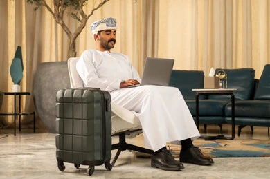 العمل عبر الحاسب المحمول، الجلوس في إحدى الفنادق الراقية، استخدام التقنيات الحديثة والمتطورة، رجل عربي خليجي عماني يرتدي الدشداشة و العمامة يجلس في لوبي الفندق بجانبه حقيبة السفر ويستخدم اللابتوب