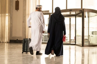 The concept of tourism and travel, spending enjoyable times during vacation, a picture from behind of an Arab Gulf Omani man wearing a dishdasha and a turban holding a travel bag in his hands walking inside the hotel lobby accompanied by his wife. The concept of tourism and travel, spending enjoyable times during vacation, a picture from behind of an Arab Gulf Omani man wearing a dishdasha and a turban holding a travel bag in his hands walking inside the hotel lobby accompanied by his wife.