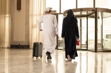 قضاء وقت عائلي سعيد في الفندق ، صورة من الخلف لرجل عربي خليجي عماني يرتدي الدشداشة و العمامة يمسك بيديه حقيبه السفر يمشي داخل بهو الفندق برفقة زوجته, رحلة عائلية سعيدة