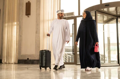 Spending an enjoyable summer trip, an Arab Gulf Omani man wearing a dishdasha and a turban is pulling a suitcase inside the hotel with his wife, expressions and gestures of happiness and joy, a family having a good time, preparing to travel on a sightseeing tour, the concept of tourism during the summer vacation.