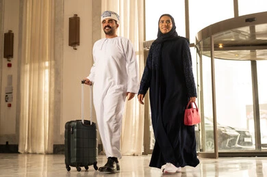 Preparing for a trip on a tourist tour, spending an enjoyable summer vacation, an Arab Gulf Omani man wearing a dishdasha and turban pulling a suitcase inside the hotel with his wife, expressions and gestures of happiness and joy, a family spending quality time, the concept of tourism during the summer holiday.