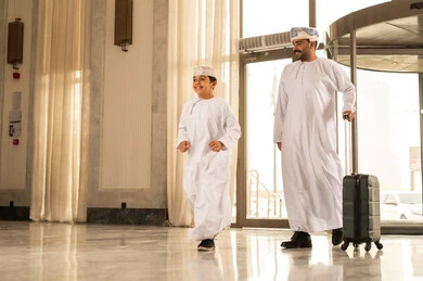 Spending an enjoyable summer trip, preparing to travel on a sightseeing tour, an Arab Gulf Omani father wearing a dishdasha and turban pulling luggage in the hotel lobby accompanied by his son, gestures of happiness and joy, the concept of tourism during the summer vacation.