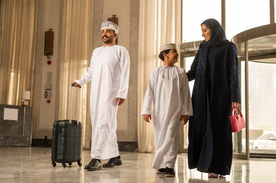 رحلة عائلية ترفيهية، ايماءات السعادة و الحماس، رجل عربي خليجي عماني يرتدي الدشداشة و العمامة يمسك بيديه حقيبة السفر يمشي داخل بهو الفندق برفقة زوجته و ابنه ، قضاء أجمل الأوقات في أفضل الفنادق الخليجية، أجواء عائلية مرحة، عطلة نهاية الأسبوع