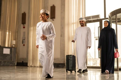 ايماءات السعادة و الحماس، رجل عربي خليجي عماني يرتدي الدشداشة و العمامة يمسك بيديه حقيبة السفر يمشي داخل بهو الفندق برفقة زوجته و ابنه ، قضاء أجمل الأوقات في أفضل الفنادق الخليجية، رحلة عائلية ترفيهية، أجواء عائلية مرحة، عطلة نهاية الأسبوع