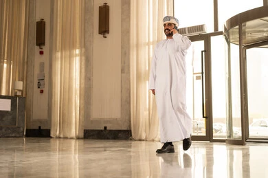 Laughing and chatting with friends and relatives, making a voice call via mobile, an Arab Omani Gulf man wearing a dishdasha and a turban walking inside the hotel holding a mobile phone in his hand.