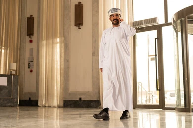 Making a voice phone call via mobile, an Arab Omani Gulf man wearing a dishdasha and a turban walks inside the hotel holding a mobile phone in his hand, laughing and chatting with friends and relatives.