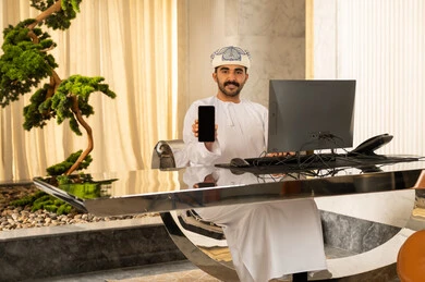 جودة تطبيقات وخدمات الهاتف المحمول، عرض شاشة سوداء فارغة، استخدام التقنيات الحديثة، رجل عربي خليجي عماني يرتدي الدشداشة و العمامة يجلس امام الحاسوب , النظر الى الكاميرا بايماءات السعادة و السرور، استقبال الضيوف وإتمام الحجزر