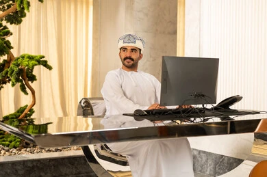 An Arab Gulf Omani man wearing a dishdasha and a turban is working on a computer, looking at something, working to increase customer satisfaction, attracting and drawing in more customers, answering inquiries and questions.