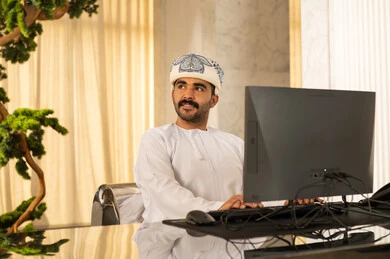 Looking at something, working to increase customer satisfaction, attracting and recruiting more customers, answering inquiries and questions, an Arab Gulf Omani man wearing a dishdasha and a turban working on a computer.