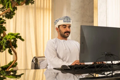 Attracting and recruiting more customers, working to increase customer satisfaction, answering inquiries and questions, an Arab Gulf Omani man wearing a dishdasha and turban working on a computer.