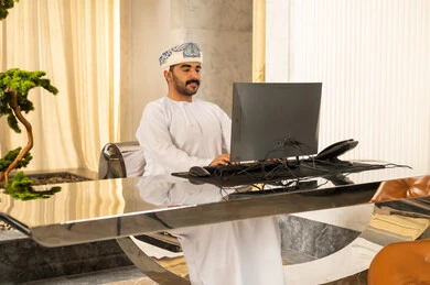 Working to increase customer satisfaction, attracting and recruiting more customers, answering inquiries and questions, an Arab Gulf Omani man wearing a dishdasha and turban working on a computer.