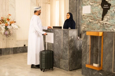 استقبال الضيوف وإتمام الحجز، مفهوم السياحه والفندقه والسفر، رجل عربي خليجي عماني يرتدي الدشداشة و العمامة يمسك حقيبة السفر و يقوم بالاستفسار فى خدمة العملاء، حسن الاستقبال والبشاشة