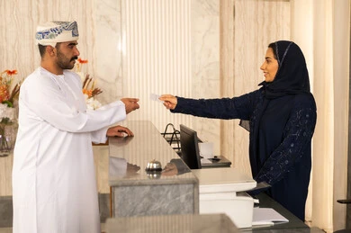 خدمة العملاء في الفندق السياحي ، موظفة استقبال عربية خليجية عمانية محجبة تقوم بتسليم بطاقة الغرفة الفندقية لسائح عربي بسعادة ، إجراءات الحجز و الدخول لدى مكتب الاستقبال في الفندق، إستلام بطاقة الغرفة ،مفهوم السياحة و الفندقة