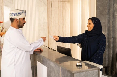 موظفة استقبال عربية خليجية عمانية محجبة تقوم بتسليم بطاقة الغرفة الفندقية لسائح عربي بسعادة ، إجراءات الحجز و الدخول لدى مكتب الاستقبال في الفندق ، خدمة العملاء في الفندق السياحي ، إستلام بطاقة الغرفة ،مفهوم السياحة و الفندقة