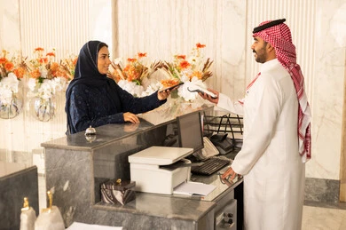 إستعلام و استفسار فى خدمة العملاء ،إجراءات الحجز والدخول لدى مكتب الاستقبال في الفندق، سياحه وفندقه وسفر، امراه عربيه خليجيه سعودية محجبة ترتدي العباءة و الحجاب تقوم بالدفع بطريقة الكترونية بإستخدام الهاتف المحمول في مكتب الاستقبال
