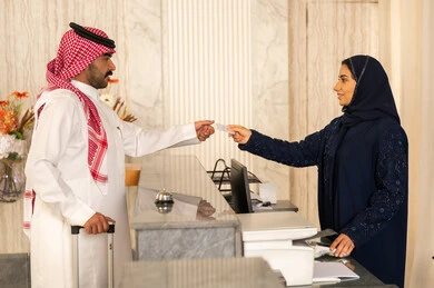حجز غرفة فندقيه عند مكتب الاستقبال، خدمة ومكتب إستقبال العملاء ،  ايماءات السرور والسعادة ، رجل عربي سعودي يرتدي الثوب الابيض و الشماغ يستلم بطاقة الغرفه