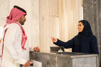 خدمة ومكتب إستقبال العملاء ،  ايماءات السرور والسعادة ، حجز غرفة فندقيه عند مكتب الاستقبال ، رجل عربي سعودي يرتدي الثوب الابيض و الشماغ يستلم بطاقة الغرفه