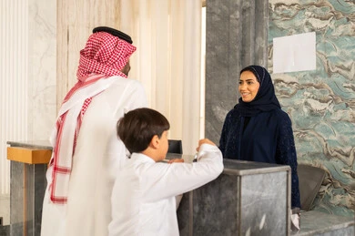 اتمام إجراءات الحجز والدخول لدى مكتب استقبال العملاء في الفندق،  مفهوم السياحه والفندقه والسفر، رجل عربي خليجي سعودي يرتدي الثوب الابيض و الشماغ يقف برفقة ابنة يقوم بالاستفسار فى خدمة العملاء