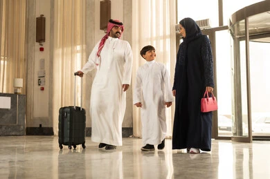 الاستمتاع في قضاء الوقت مع الأهل، الاستعداد لقضاء رحلة سياحية ممتعه مع العائلة, تبادل الاحاديث بتعابير السرور و السعادة, رجل عربي خليجي سعوجي يرتدي الثوب الابيض و الشماغ يمسك بيده حقيبة السفر برفقة زوجته و ابنه