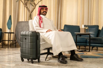 النظر الى الكاميرا بتعابير السعادة وا لسرور، قضاء رحلة صيفية ممتعة، القيام بجولة ترفيهية، مفهوم السياحة في إجازة الصيف، رجل عربي خليجي سعودي يرتدي الثوب الابيض و الشماغ يجلس بجانب حقيبة السفر ، السياحة في إجازة الصيف