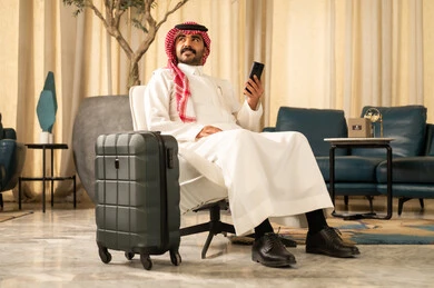 Communication and browsing the internet through social media, an Arab Gulf Saudi man wearing a white thobe and a shemagh sitting next to a suitcase holding a mobile phone in his hands, spending enjoyable times playing online games on his mobile phone, looking at something.