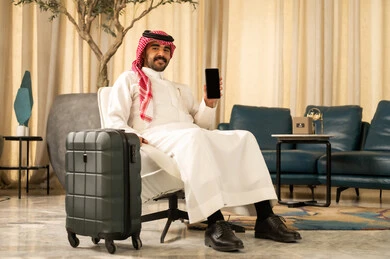 النظر الى الكاميرا بتعابير السعادة و السرور، الاستفادة من خدمات وتطبيقات الجوال، دمج التكنولوجيا والاجهزة الحديثة في الحياة اليومية، رجل عربي خليجي سعودي يرتدي الثوب الابيض و الشماغ يجلس بجانب حقيبة السفر يقوم بعرض شاشة سوداء فارغة