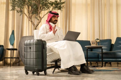 Using a laptop during waiting time, making a video call, raising a hand in a gesture of greeting and peace, chatting with family and friends, getting ready to go on an enjoyable trip, an Arab Gulf Saudi man wearing a white thobe and a ghutrah sitting on a chair with a suitcase beside him.