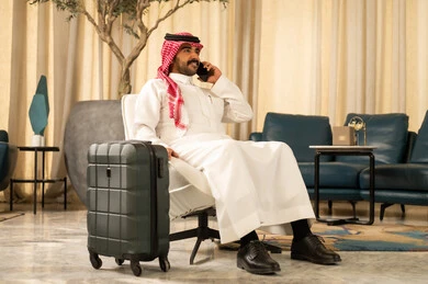 إجراء مكالمة هاتفية عبر الجوال, التواصل مع الأهل والأصدقاء,رجل  عربي سعودي خليجي يرتدي الثوب الابيض و الشماغ يجلس بجانب حقيبة السفر يمسك بيده الهاتف المحمول , حجز مباشر عبر تطبيقات وبرامج السياحة