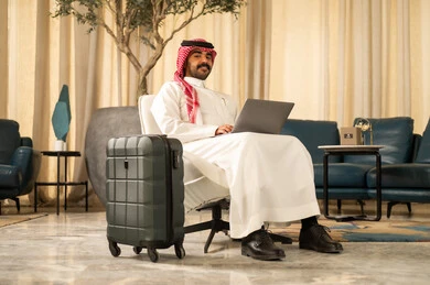 ادراة شؤون العمل عن بعد، استخدام جهاز تقني حديث و متطور , التجهيز للسفر والرحلات الممتعة, رجل عربي خليجي سعودي يرتدي الثوب التقليدي والشماغ يجلس بجانب حقيبة السفر يمسك بيده الحاسب المحمول