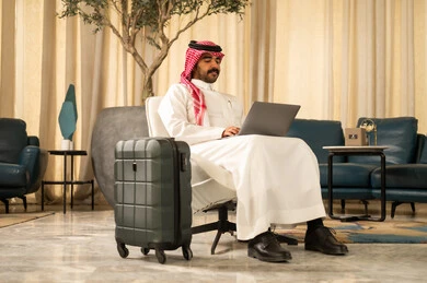 استخدام جهاز تقني حديث و متطور , التجهيز للسفر والرحلات الممتعة, رجل عربي خليجي سعودي يرتدي الثوب التقليدي والشماغ يجلس بجانب حقيبة السفر يمسك بيده الحاسب المحمول , ادراة شؤون العمل عن بعد