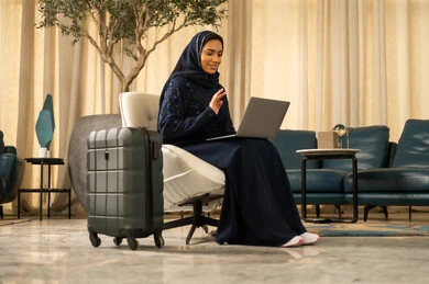 اجراء مكالمة فيديو، رفع كف اليد بتعبير التحية و السلام، الدردشة مع الأهل والأصدقاء، الاستعداد للذهاب في رحلة ممتعة، استخدام الحاسوب المحمول في وقت الانتظار، امراة عربية خليجية سعودية ترتدي العباءة  تجلس على كرسي وبجانبها حقيبة سفر