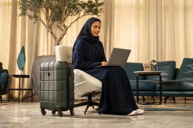 Browsing a website and internet application, chatting with family and friends, preparing to go on enjoyable trips, using a laptop while waiting, an Arab Gulf Saudi Muslim woman wearing an abaya sitting on a chair with a suitcase beside her.