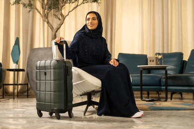 قضاء رحلة صيفية ممتعة, الاستعداد لبدء العطلة خارج البلاد, السياحة في إجازة الصيف, امراة عربية خليجية سعودية محجبة ترتدي العباءة تجلس على كرسي تضع يدها على حقيبة السفر تنظر إلى الاعلى بإيماءات التأمل والسرور