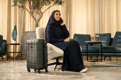 الإجابة والرد على الاتصالات باستخدام الهاتف المحمول، الشعور بالسعادة والفرح، السياحة في إجازة الصيف, الاستعداد لقضاء رحلة صيفية ممتعة, امراة عربية خليجية سعودية محجبة ترتدي العباءة تجلس بجانب حقيبة السفر, التواصل والاطمئنان على الأصدقاء