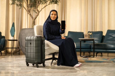 قضاء رحلة ممتعة خارج البلاد, استخدام جهاز تقني حديث متطور,امراة عربية خليجية سعودية محجبة ترتدي العباءة بجانبها حقيبة سفر تمسك بيدها جوال , عرض شاشة سوداء فارغة عبر الهاتف المحمول
