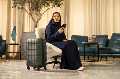 Browsing social media, using a modern and advanced technical device, preparing for travel and enjoyable trips, an Arab Gulf Saudi woman wearing a hijab and an abaya is sitting on a chair in the hotel next to her travel bag holding a mobile phone.