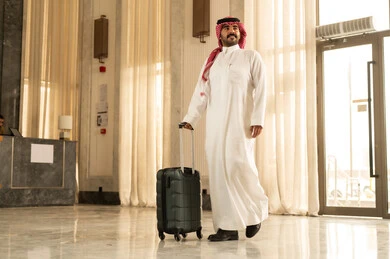 Tourism during the summer vacation, the feeling of happiness and joy, preparing for an enjoyable summer trip, an Arab Gulf Saudi man wearing a white thobe and a shemagh stands inside the hotel holding a suitcase with gestures of excitement.
