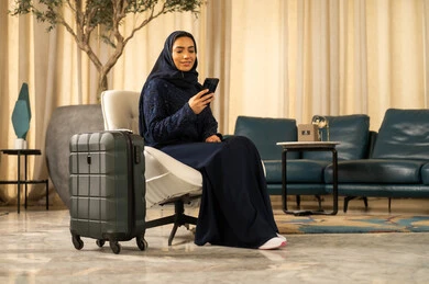 استخدام جهاز تقني حديث و متطور , التجهيز للسفر والرحلات الممتعة,امراة عربية خليجية سعودية محجبة ترتدي العباءة تجلس على كرسي فى الفندق بجانبها بيديها حقيبة السفر تمسك الهاتف المحمول، تصفح موقع التواصل الاجتماعي
