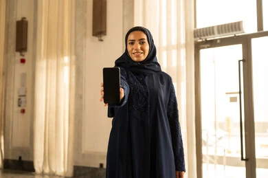 The use of modern and advanced devices and technology, looking at the camera with expressions of happiness and joy, an Arab Gulf Saudi woman wearing a hijab and an abaya standing inside a tourist hotel and displaying a blank black screen on her mobile phone.