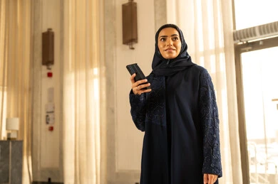 Online shopping, browsing social media, a Saudi Arabian Gulf woman wearing a hijab and an abaya stands inside a tourist hotel holding a mobile phone in her hand, looking at something with gestures of joy.