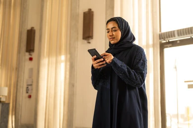 Spending enjoyable time on the mobile, browsing social media, an Arab Gulf Saudi woman in a hijab wearing an abaya stands inside a tourist hotel holding a mobile phone in her hand, looking with gestures of joy.