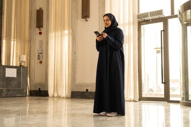Browsing social media, having a good time on the mobile, an Arab Gulf Saudi woman wearing a hijab and an abaya stands inside a tourist hotel holding a mobile phone in her hand, looking with gestures of joy.