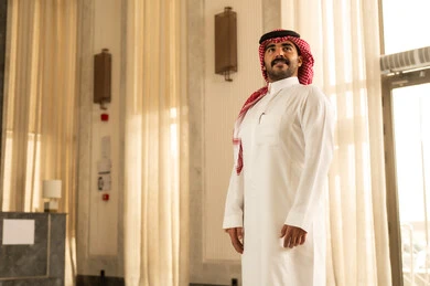 A Saudi Arabian Gulf man wearing a white thobe and a ghutrah stands inside a hotel, looking at something with gestures of joy and happiness, showing interest and elegance in his appearance, embodying the concepts of masculinity, strength, and self-confidence.