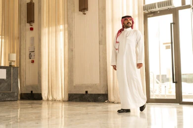 الاهتمام والأناقه في المظهر الخارجي، مفهوم الرجولة والقوة والثقة بالنفس، رجل عربي خليجي سعودي يرتدي الثوب الابيض و الشماغ يقف داخل الفندق ينظر إلى شيء ما بإيماءات السرور والفرح