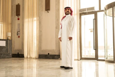 مفهوم الرجولة والقوة والثقة بالنفس، رجل عربي خليجي سعودي يرتدي الثوب الابيض و الشماغ يقف داخل الفندق ينظر إلى شيء ما بإيماءات السرور والفرح، الاهتمام والأناقه في المظهر الخارجي