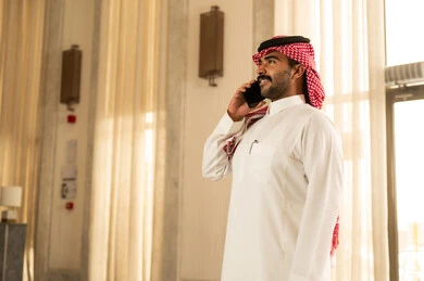 التواصل مع الاصدقاء، اجراء مكالمة هاتفية، رجل عربي خليجي سعودي يرتدي الثوب الابيض و الشماغ يقف داخل الفندق يستخدم الهاتف المحمول، تعابير السعادة و السرور
