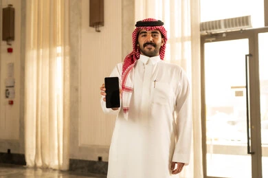 The concept of technological and technical developments, an Arab Gulf Saudi man wearing a white thobe and a shemagh stands inside a hotel displaying a blank black screen via a mobile phone, keeping up with modern contemporary progress.