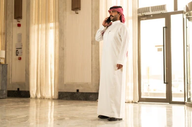 Making a phone call, a Saudi Arabian Gulf man wearing a white thobe and a ghutrah stands inside a hotel using a mobile phone, communicating with friends, expressions of happiness and joy. Making a phone call, a Saudi Arabian Gulf man wearing a white thobe and a ghutrah stands inside a hotel using a mobile phone, communicating with friends, expressions of happiness and joy.