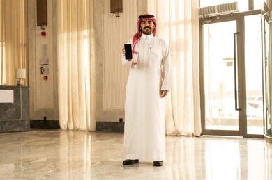 مواكبة التقدم العصري الحديث، مفهوم التطورات التقنية والتكنولوجية، رجل عربي خليجي سعودي يرتدي الثوب الابيض و الشماغ يقف داخل الفندق يقوم بعرض شاشة سوداء فارغة عبر الهاتف المحمول